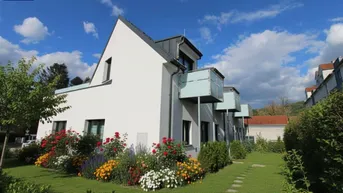 Moderne Reihenhäuser mit gepflegtem Vorgarten, bunten Blumenbeeten und Balkonen unter blauem Himmel.