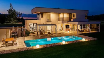 Moderne Villa mit beleuchtetem Pool, Garten und gemütlichem Essbereich im Freien bei Dämmerung.