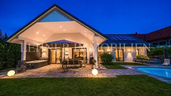 Elegantes Haus bei Nacht mit beleuchteter Terrasse, Pool und Whirlpool, umgeben von Grün.