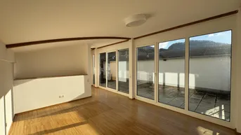Lichtdurchfluteter Raum mit Holzboden, Dachschräge und breiter Fensterfront zur Terrasse.