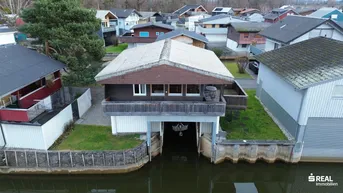 Ansicht des Hauses mit Bootsgarage direkt am Wasser und umliegender Grünfläche.