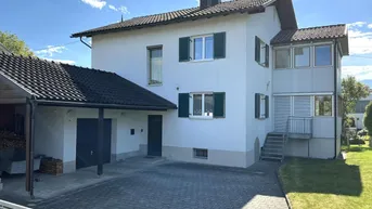 Weißes Einfamilienhaus mit Carport und gepflasterter Einfahrt unter blauem Himmel.