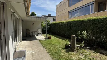 Sonniger Außenbereich mit gepflasterter Terrasse, Rasenfläche und Hecke.