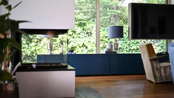 Helles Wohnzimmer mit modernem Kamin, Holzboden und großem Fenster mit Blick ins Grüne.