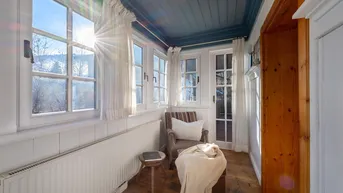 Heller Wintergarten mit vielen Fenstern, Holzboden und einem gemütlichen Sessel.