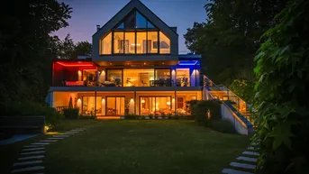 Moderne Villa bei Nacht mit beleuchteten Terrassen und großzügiger Gartenanlage am Seeufer.