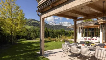 Überdachte Terrasse mit Esstisch und Stühlen, umgeben von einem grünen Garten und Bergblick.