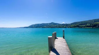 Blick vom Ende des Holzstegs auf das weite, türkisfarbene Seewasser und die bewaldeten Berge.