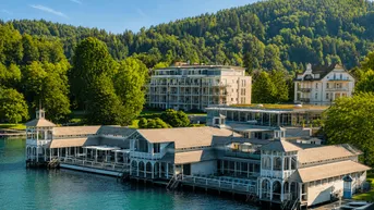 Panoramablick auf moderne Gebäude am Seeufer mit grünen Hügeln im Hintergrund.
