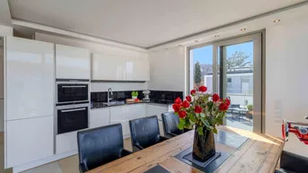 Moderne Küche mit weißen Hochglanzfronten und Essbereich, ausgestattet mit einem Holztisch und Blick ins Freie.