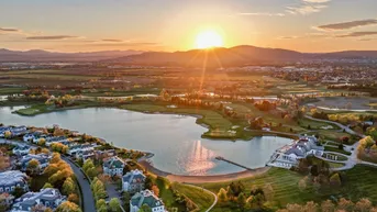 Weitläufige Luftaufnahme der Wohnanlage am Seeufer bei Sonnenuntergang mit Golfplatz und Bergen im Hintergrund.