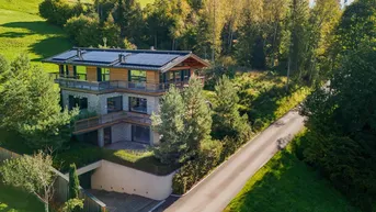 Luftaufnahme eines modernen Chalets mit Photovoltaikanlage auf dem Dach, umgeben von Wald und Wiesen.