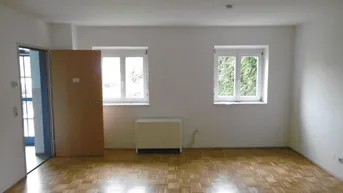 Helles Wohnzimmer mit Parkettboden und zwei Fenstern, die einen Blick ins Grüne bieten.