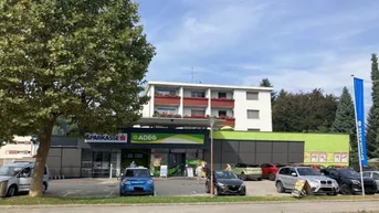 Gebäude mit Supermarkt im Erdgeschoss und Wohnungen darüber, umgeben von einem Parkplatz.