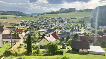 Panoramablick auf ein malerisches Dorf in einer grünen Hügellandschaft unter bewölktem Himmel.