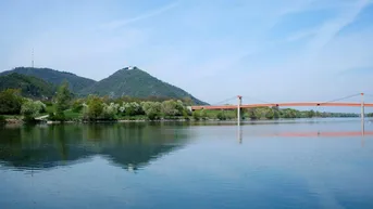 Weitläufiger Blick auf einen ruhigen See mit Bergen und einer markanten Brücke im Hintergrund.