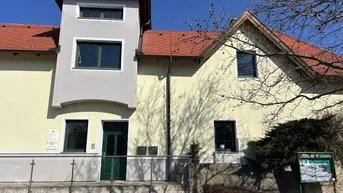 Gelbe Fassade eines Gebäudes mit grauem Anbau und rotem Ziegeldach unter blauem Himmel.