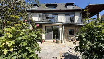 Rückansicht des Hauses mit großer Steinterrasse und darüberliegendem Balkon, umgeben von Pflanzen.