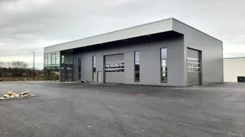 Modernes Gewerbegebäude mit grauer Fassade, großen Fenstern und einem weitläufigen Parkplatz unter bewölktem Himmel.