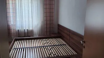 Helles Schlafzimmer mit Holzbettrahmen, Parkettboden und Vorhängen am Fenster.