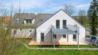 Modernes Haus mit großer Holzterrasse und Balkon, umgeben von grüner Landschaft unter blauem Himmel.