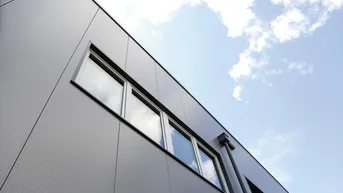 Moderne Fassade eines Gebäudes mit grauen Metallpaneelen und horizontalen Fenstern unter blauem Himmel.