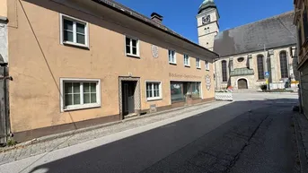 Historisches Gebäude an einer Straße mit einer Kirche im Hintergrund unter blauem Himmel.