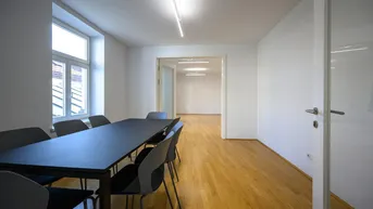 Heller Konferenzraum mit Holzboden und großem Tisch, der zu einem weiteren Raum führt.