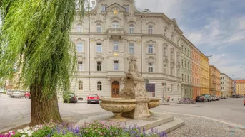 Historisches Gebäude mit aufwendiger Fassade und einem Brunnen im Vordergrund an einer belebten Straße.