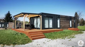 Modernes Modulhaus mit Holzterrasse und Pergola, umgeben von grüner Landschaft.