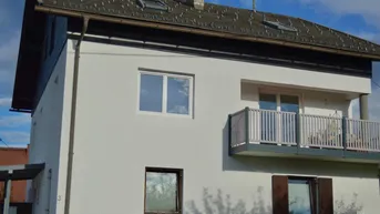 Ansicht der weißen Hausfassade mit Balkon und dunklem Dach unter blauem Himmel.
