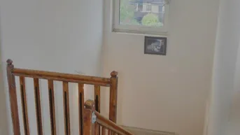 Heller Treppenaufgang mit Holzgeländer und Fenster, das einen Blick ins Grüne bietet.