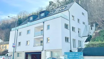 Modernes Mehrfamilienhaus mit weißer Fassade, Balkonen und Dachgauben in grüner Hanglage.