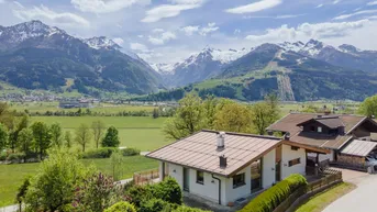 Idyllisches Haus mit Bergblick, umgeben von grüner Landschaft und Bäumen unter blauem Himmel.