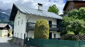 Traditionelles Haus mit Balkon und Satteldach, umgeben von grüner Natur und Bergblick.