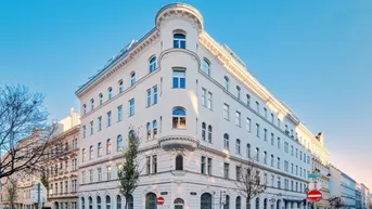 Weißes Eckgebäude mit klassischer Architektur und vielen Fenstern in städtischer Umgebung.