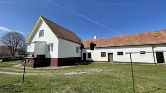 Ländliches Anwesen mit weiß getünchtem Haus, braunem Sockel und großem Rasenbereich unter blauem Himmel.