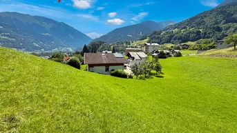 Panoramablick auf Häuser in einer grünen Berglandschaft mit sonnigem Himmel.