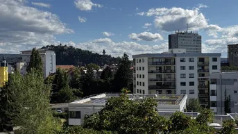 Stadtansicht mit Wohngebäuden, grünen Bäumen und bewölktem Himmel über den Hügeln.