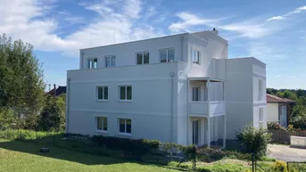 Modernes Mehrfamilienhaus mit weißer Fassade, Balkonen und umliegender Grünfläche unter blauem Himmel.