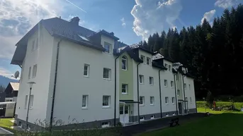 Mehrfamilienhaus mit heller Fassade und Satteldach, umgeben von Grünflächen und Wald.