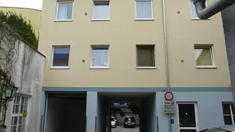 Mehrstöckiges Gebäude mit beiger Fassade, blauer Sockelzone, Garageneinfahrt und Durchgang zum Hinterhof.