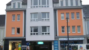 Mehrstöckiges Gebäude mit orangefarbener und grauer Fassade, großen Fenstern und Geschäften im Erdgeschoss.