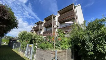 Mehrstöckiges Gebäude mit Holzbalkonen und üppigem Garten im Vordergrund unter blauem Himmel.