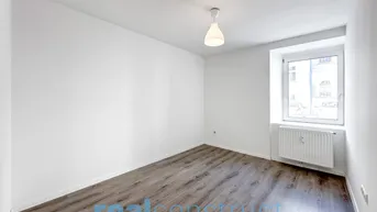 Helles, leeres Zimmer mit Laminatboden und einem Fenster, das viel Tageslicht hereinlässt.