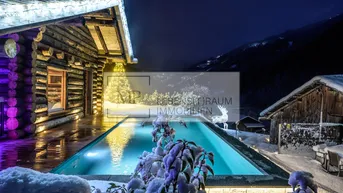 Beleuchteter Außenpool im Schnee vor einer rustikalen Holzhütte mit winterlicher Bergkulisse bei Nacht.