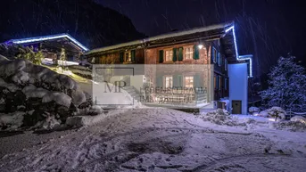 Charmantes Holzchalet in verschneiter Berglandschaft bei Nacht, mit beleuchteter Fassade und Zufahrt.