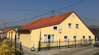 Gelbes Einfamilienhaus mit rotem Ziegeldach und umzäuntem Vorgarten unter blauem Himmel.