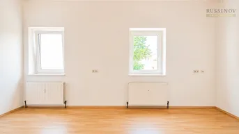 Geräumiger, heller Raum mit Holzdeckenverkleidung, Laminatboden und zwei Fenstern mit Grünblick.