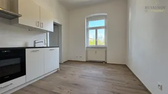 Heller Raum mit integrierter Küchenzeile, Laminatboden und Fenster mit Blick ins Grüne.